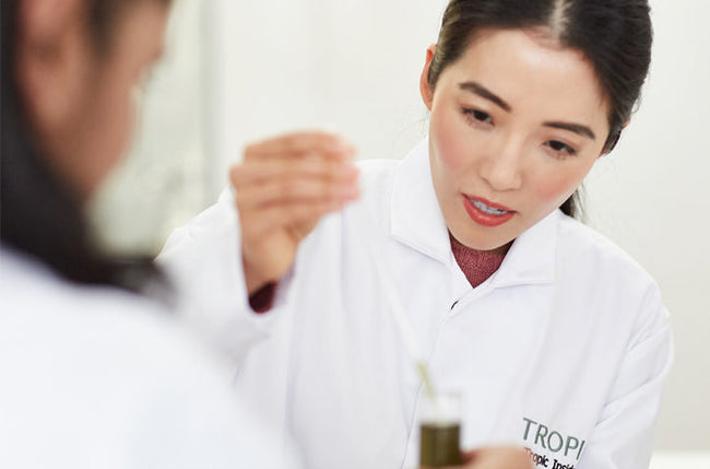 This image shows our founder Susie in a lab coat using a pipette to sample liquid from a test tube with another member of staff opposite her.